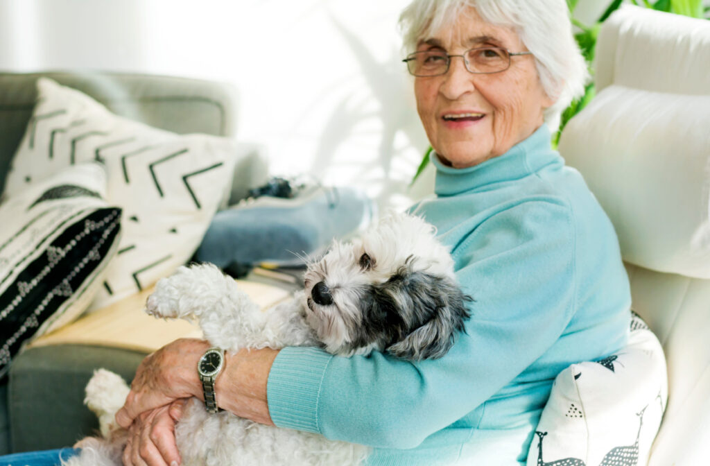 A senior woman smiling and sitting on a couch hugging her dog while looking directly at the camera