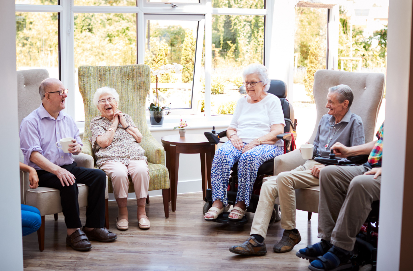 A group of older adults sitting in a semicircle in a common area, drinking tea or coffee and laughing
