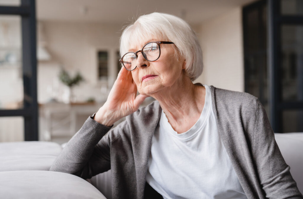 An older adult woman with glasses on sitting on a couch and looking out the window with a serious expression