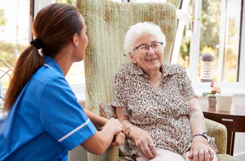 An older adult woman in a memory care facility sitting on a chair smiling and having a conversation with a nurse.