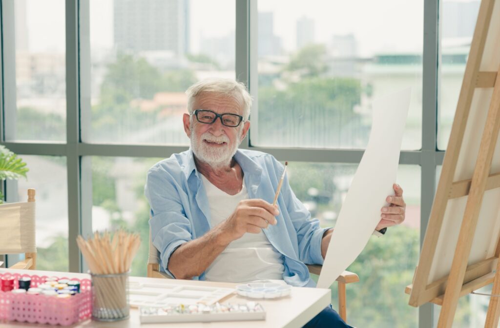 A smiling older adult man sitting in an art studio and holding a paintbrush in one hand and a piece of paper in the other hand.